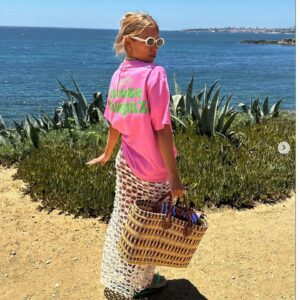 Foto de uma mulher com blusa rosa chiclete, óculos de sol branco, saia com furos e bolsa de palha em frente ao mar. Foto demonstra a moda portuguesa.