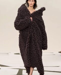 Foto de modelo com um sobretudo marrom com tem textura diferenciada, uma das tendências da moda no ano. A textura do tecido parecem caichos grandes.