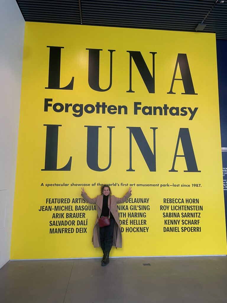 Foto da Silvia Sigliano na entrada da exposição Luna Luna: Forgotten Fantasy. Atrás da Silvia há uma parede amarela com o nome da exposição grande em preto e o nome dos artistas.