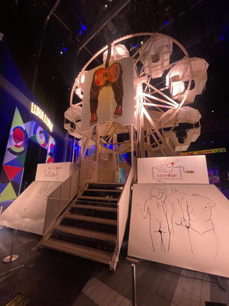 Visão de traz da roda gigante. A estrutura possui várias obras de arte com desenhos de corpos e outros elementos a mão.