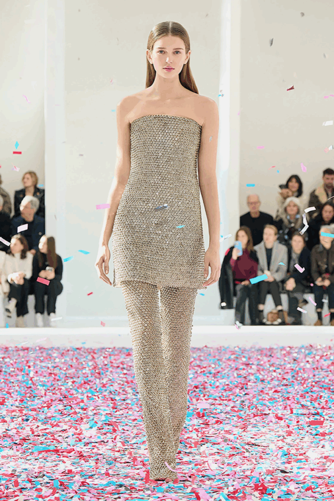 Modelo com roupa da Courreges. As peças contém dois dos shapes de moda mais vistos: vestido com ombros de fora por cima de uma calça. Ambos são feitos de um tecido com textura brilhosa.