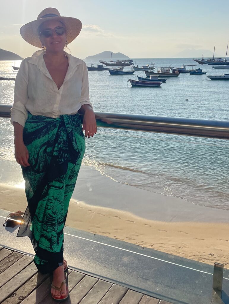 Foto da Silvia Scigliano em um deck à beira do mar usando uma camisa de linho, uma canga longa verde amarrada na cintura e um chapeu de palha.