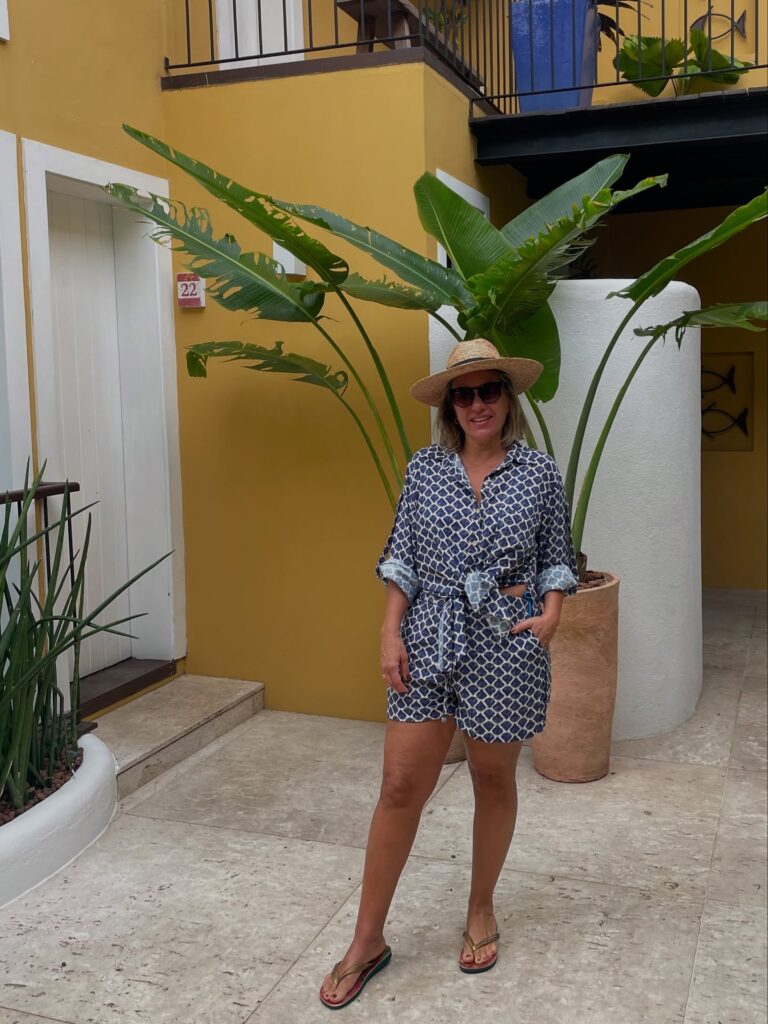 Foto da Silvia usando camisa de manga com amarração na cintura e shorts estampado. Também usa sandalha rasteira e chapeu de palha.