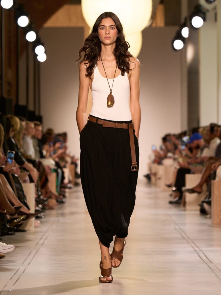Modelo branca usa calça Aladin preta, regata banca e acessários marrons em desfile da marca Michael Kors.