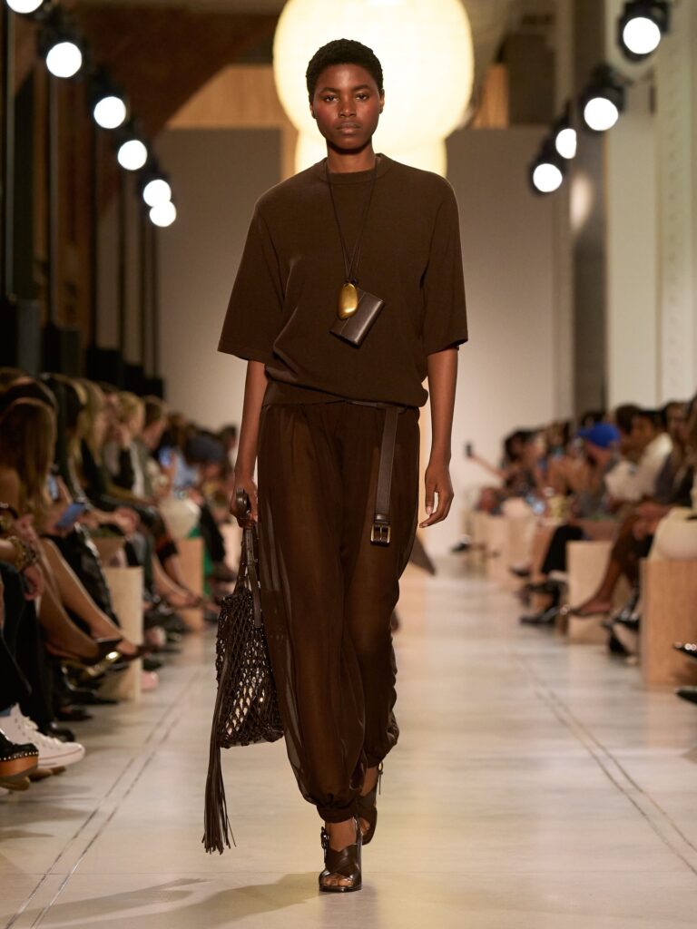 Modelo negra usa conjunto marrom em desfile de Michael Kors, composto por blusa de mangas langas, calça aladin translúcida, bolsa vasada e sandalha de tiras.