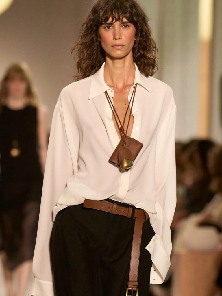 Modelo usa camisa branca e calça preta. O destaque é para o colar marcante e cinto em couro, reforçando a tendência de acessórios.