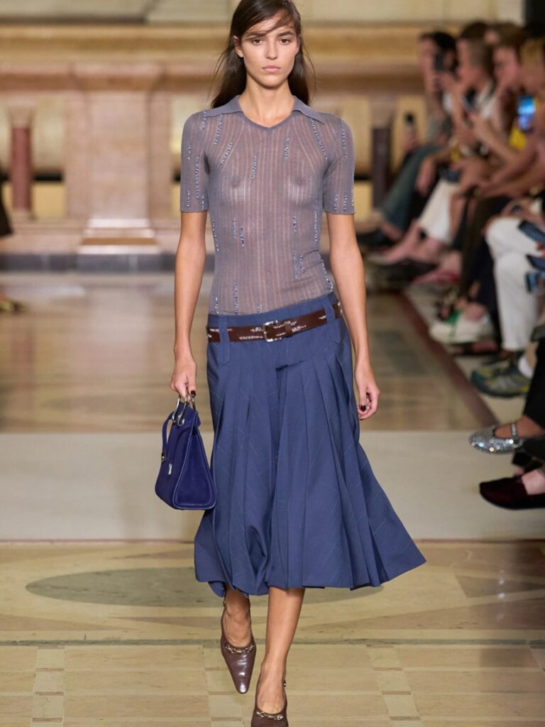 Modelo usa blusa transparente com saia de cintura baixa azul e cinto marrom que chama a atenção.