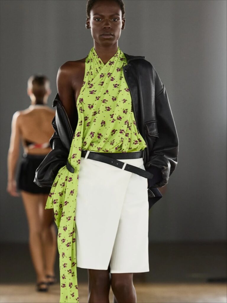 Modelo usa saia branca com cinto preto. A saia possui uma das guias para o cinto mais baixas, deixando a ponta do cinto cruzando a peça.