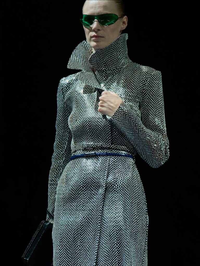 Modelo com visual futurista usando um sobretudo prata e dois cintos: um do mesmo tecido do vestido e outro azul